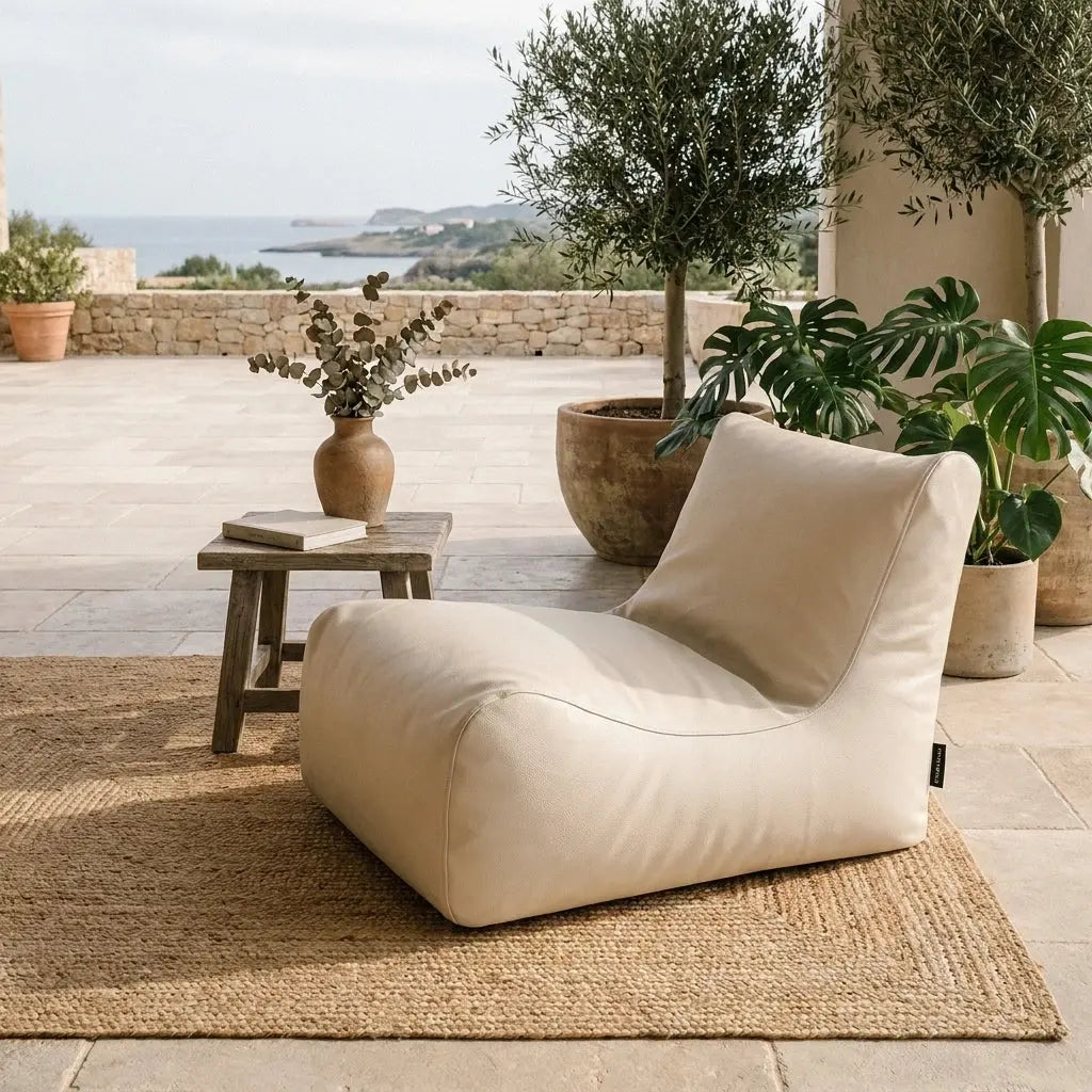 Sittsäck Lounge 100 utomhus beige bredvid ett litet bord på en terrass med växter och havsutsikt.