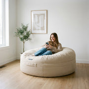A woman relaxes on a Saccosäck Roll 135 Madu Cream bean bag in a bright, minimalist living room with wood floors.