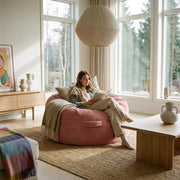 Cozy living room with a woman reading on a pink beanbag chair, featuring natural wood furniture, a jute rug, and modern decor.