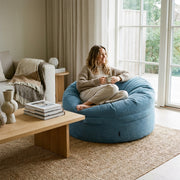 A cozy, modern living room featuring a woman relaxing in a blue corduroy bean bag chair by a bright window.